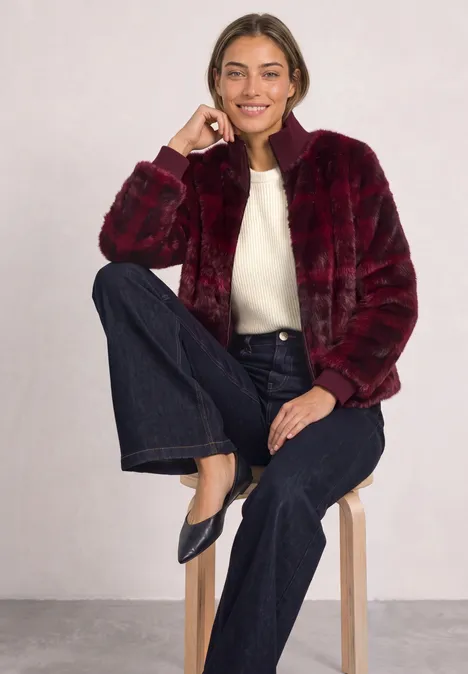 Fake Fur Blouson mit Strickdetails und Print plummy brown Frau in roter Plüschjacke, weißem Pullover und dunklen Jeans sitzt lässig auf einem Hocker vor grauem Hintergrund.