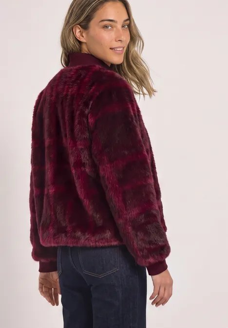 Fake Fur Blouson mit Strickdetails und Print plummy brown Frau in bordeauxroter Kunstpelz-Bomberjacke und Jeans blickt über die Schulter vor hellem Hintergrund.