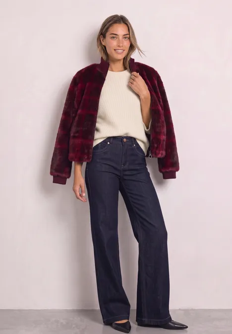 Fake Fur Blouson mit Strickdetails und Print plummy brown Frau in bordeauxroter Kunstfelljacke, cremeweißem Pullover und weiten, dunkelblauen Jeans vor hellem Hintergrund.