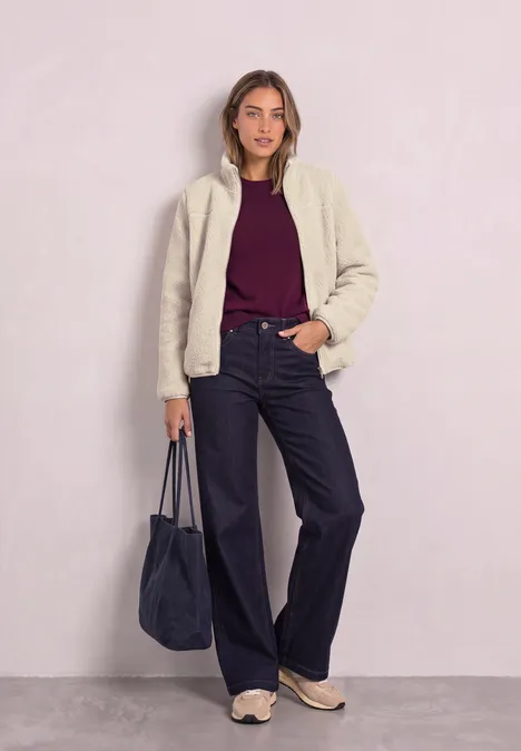 Teddy-Jacke mit Stehkragen und Zipper Soft Fawn Beige Frau im cremefarbenen Blouson, bordeauxrotem Pullover, weiten schwarzen Hosen, Sneakern und blauer Tasche.