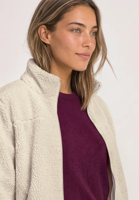 Teddy-Jacke mit Stehkragen und Zipper Soft Fawn Beige Frau trägt beigen Fleece-Mantel über lila Pullover, schaut seitlich, neutraler Hintergrund.