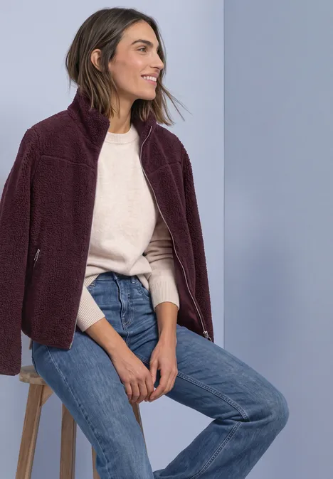 Teddy-Jacke mit Stehkragen und Zipper rustic rouge Frau sitzt lächelnd auf Hocker, trägt bordeauxfarbenes Fleece-Jacke, hellen Pullover und Jeans.
