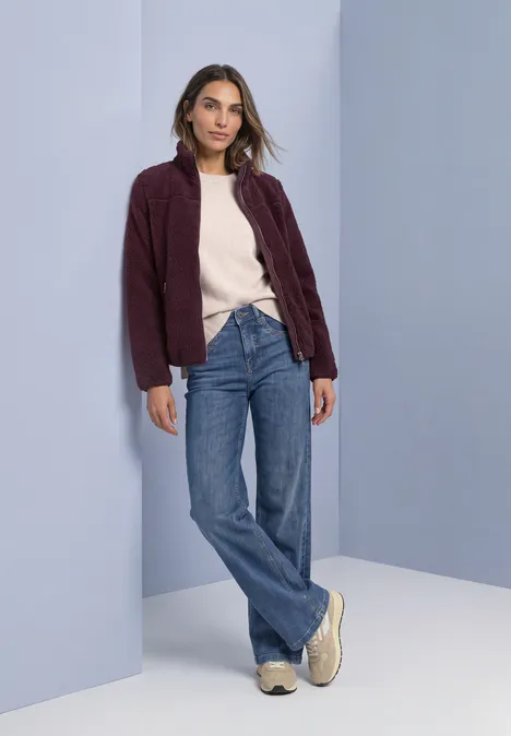 Teddy-Jacke mit Stehkragen und Zipper rustic rouge Frau lehnt lässig an Wand, trägt weinroten Cardigan, hellen Pullover, weite Jeans und beige Schuhe