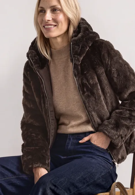Frau auf Stuhl, lächelnd in brauner Pelzjacke, beigefarbenem Pullover und blauen Jeans.