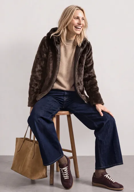 Frau sitzt lachend auf Hocker, trägt braune Kunstpelzjacke, beigen Pullover, weite Jeans, Sneaker und Tasche.