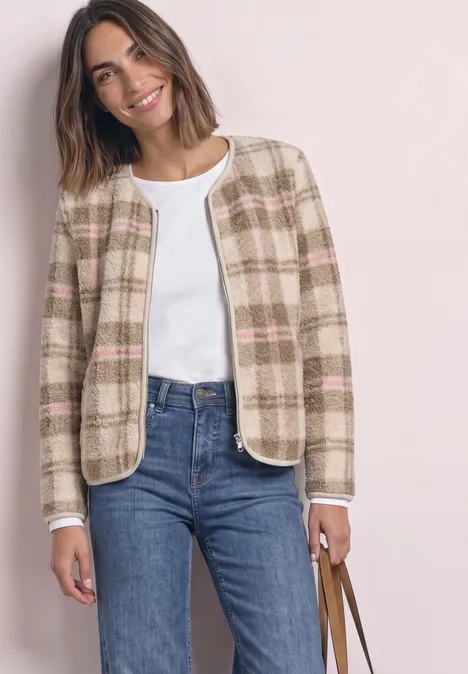 Femme souriante en veste courte à carreaux beige-rose, top blanc, jean bleu, sac marron.