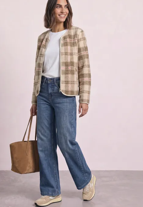 Femme en veste beige à carreaux, t-shirt blanc, jean droit, baskets beige et sac marron devant un fond clair.