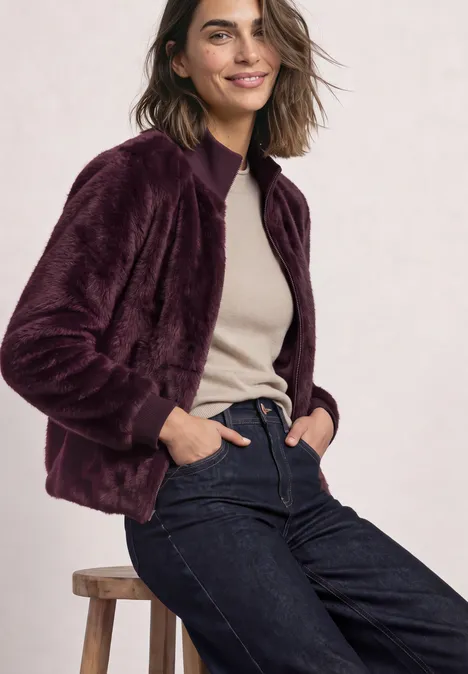 Blouson van imitatiebont met gebreide details en print rustic rouge Frau in violettem Plüschblouson, beigem Oberteil und dunkler Jeans lehnt sich lächelnd an einen Hocker.