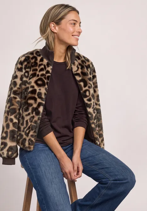 Frau sitzt auf Hocker, trägt Leo-Jacke, schwarzen Pullover und Jeans, lächelt seitlich.