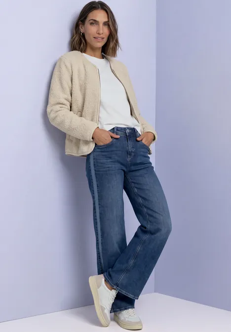 Veste en peluche à col rond et détails en velours côtelé Soft Fawn Beige Frau in beiger Teddy-Jacke, weißem T-Shirt, blauen Jeans und weißen Sneakern vor lila Wand.