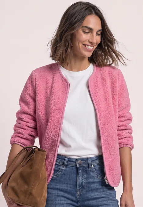 Une femme souriante porte une veste rose, un t-shirt blanc, un jean bleu et tient un sac marron.