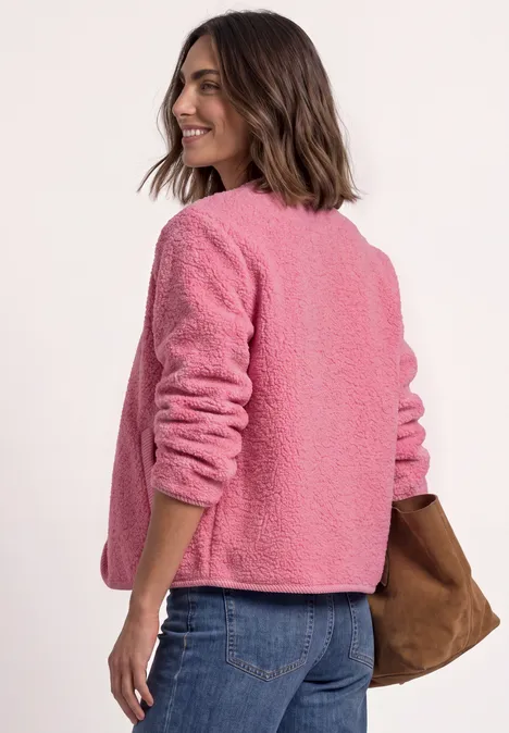 Femme en veste teddy rose, tourne le dos et regarde par-dessus l’épaule, jean et sac marron.