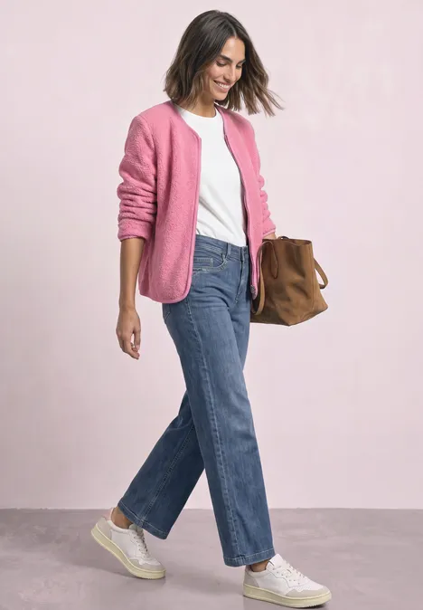 Une femme porte un T-shirt blanc, un gilet rose, un jean droit bleu, des baskets blanches et un sac marron.