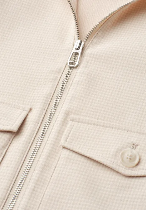 Nahaufnahme einer beigen Jacke mit Metallreißverschluss und zwei Brusttaschen mit Klappen.
