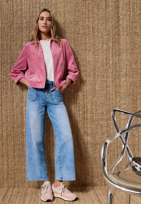 Person in rosa Jacke, weißem Top, weiten Jeans und rosa Sneakers vor strukturiertem Hintergrund.