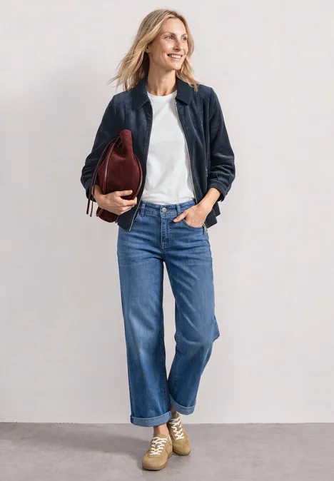 Frau trägt blaue Jeans, weißes Shirt, dunkelblaue Jacke, beige Sneaker und hält eine rote Handtasche.