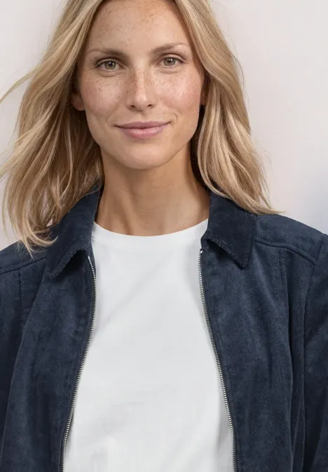 Dame mit blonden Haaren, trägt weißes T-Shirt und dunkelblaue Jacke.