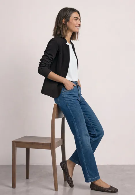 Blazer mit Reverskragen und Struktur Black Frau in legerem Outfit: schwarzer Blazer, weißes Shirt, blaue Jeans und flache Schuhe lehnt an einem Stuhl.