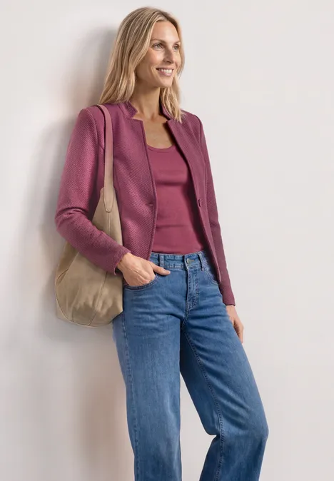 Femme portant une veste framboise, top assorti, jean bleu et sac en tissu beige devant un mur blanc.