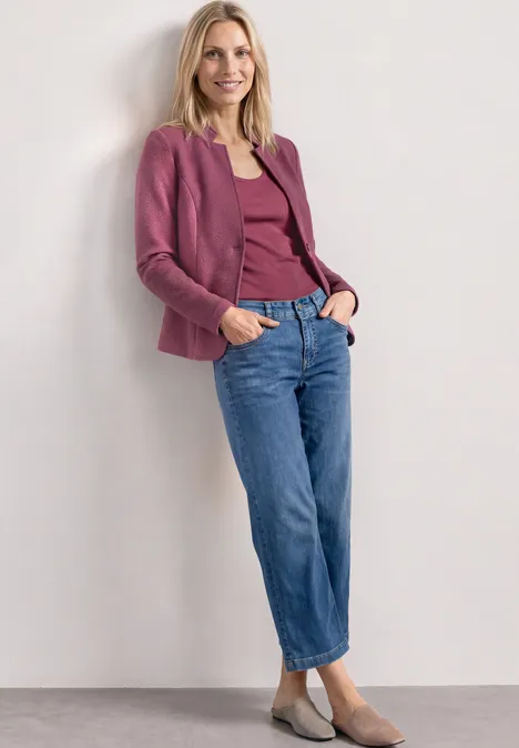 Femme appuyée sur un mur blanc, portant un blazer et un haut violets, un jean bleu et des ballerines beiges.