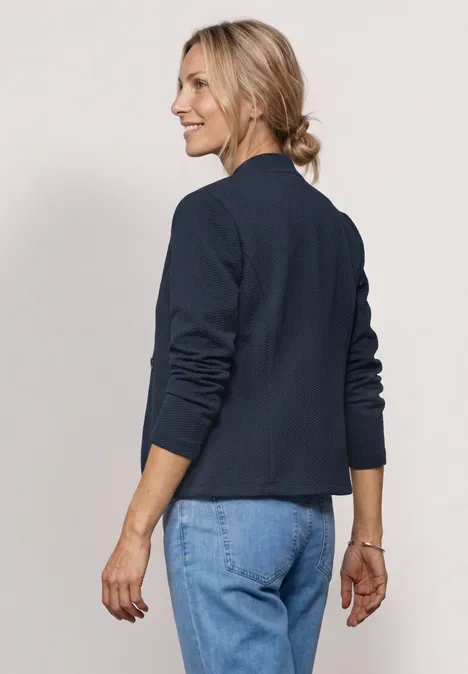 Eine Frau trägt eine dunkelblaue Jacke und Jeans, steht seitlich mit unscharfem Gesicht und Dutt.
