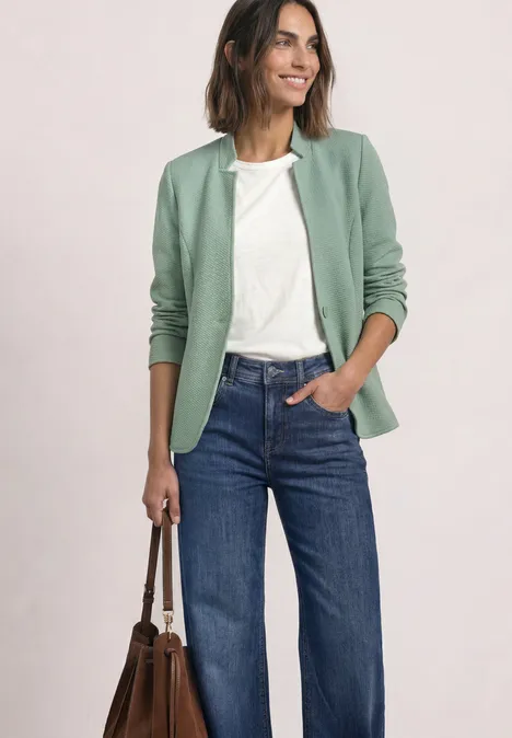 Blazer met reverskraag en structuur eucalyptus green Frau in grünem Blazer, weißem Shirt, weiten Jeans mit Handtasche, lächelnd vor neutralem Hintergrund.