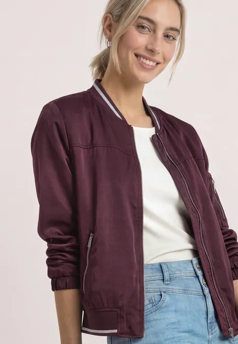 Eine Frau trägt eine lila Bomberjacke, weißes Shirt und blaue Jeans, lächelt vor hellem Hintergrund.