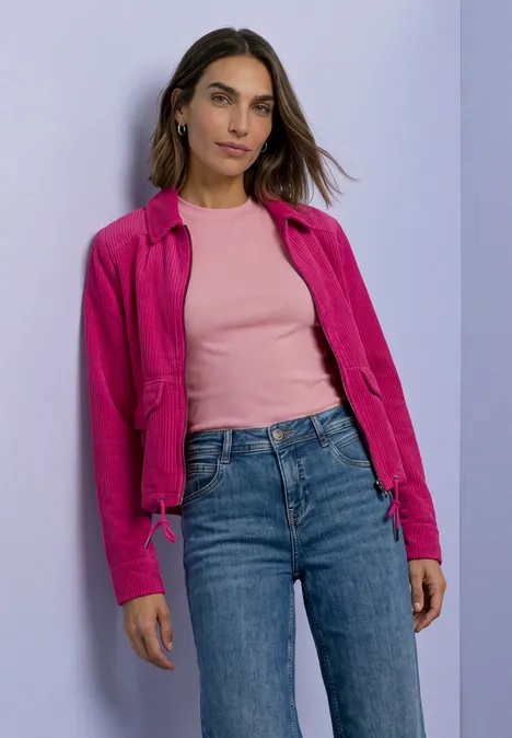 Eine Frau in pinker Jacke, rosa Top und Jeans vor lila Wand.