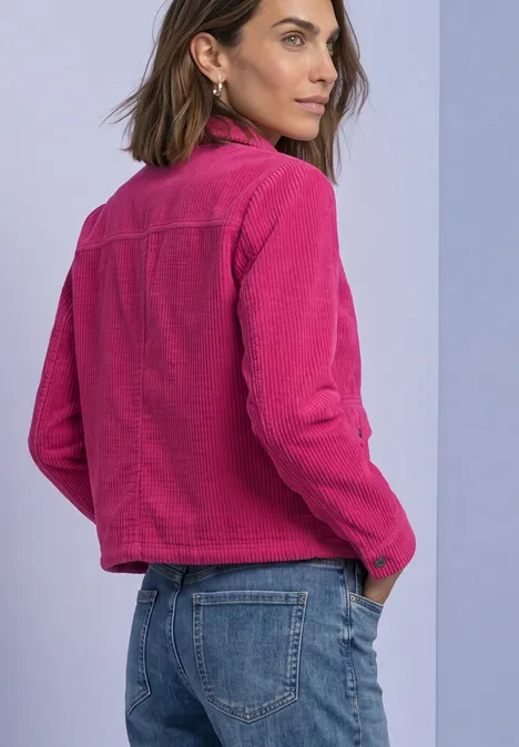Frau in rosa Jacke und blauen Jeans blickt über die Schulter vor hellem Hintergrund.