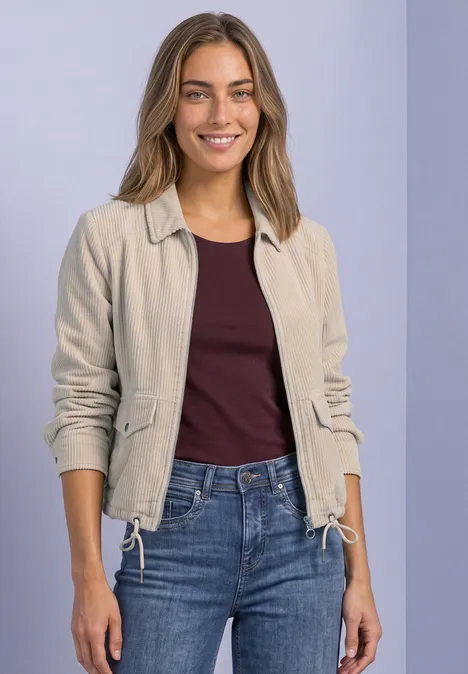 Cordjacke mit Hemdkragen und Zipper clay sand Eine lächelnde Frau in beigefarbener Jacke, bordeauxrotem Shirt und Jeans vor hellviolettem Hintergrund.