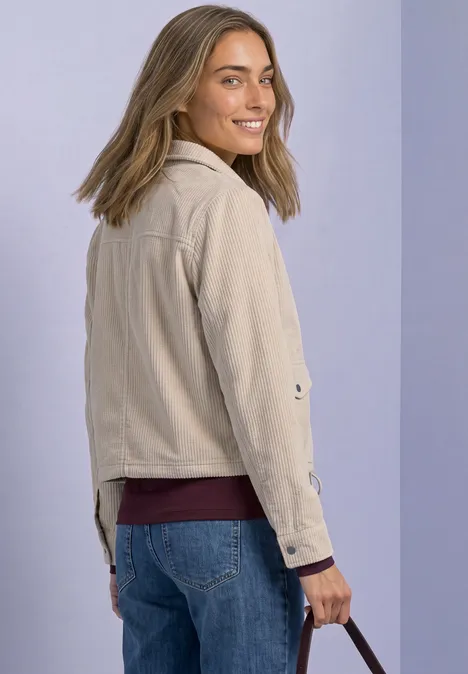 Cordjacke mit Hemdkragen und Zipper clay sand Frau im beigen Jackett, blauen Jeans und weinrotem Top, lächelt über die Schulter, trägt Tasche vor lila Hintergrund.