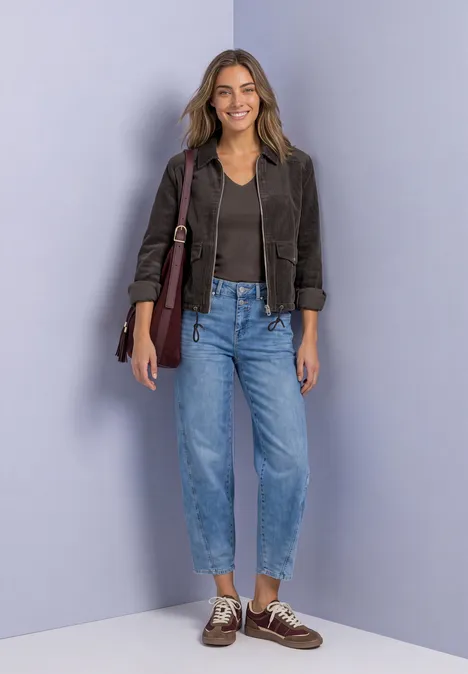 Frau in Lederjacke, schwarzem Top, Jeans und Bootsschuhen lächelt und trägt eine Umhängetasche vor fliederfarbener Wand.