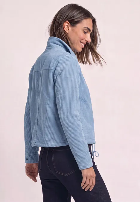 Frau mit hellblauer Cordjacke und dunklen Jeans, seitlich stehend vor neutralem Hintergrund.