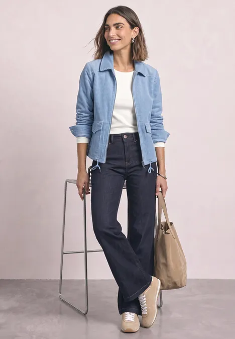 Frau in heller Jacke, weißem Top, dunklen Schlagjeans und hellen Schuhen trägt Tasche vor neutralem Hintergrund.