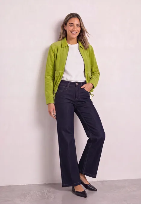 Veste en velours côtelé avec col chemise et fermeture à zip apple lime Frau lehnt an Wand, trägt grünen Blazer, weißes Shirt, dunkle weite Hose und schwarze Ballerinas, lächelnd.