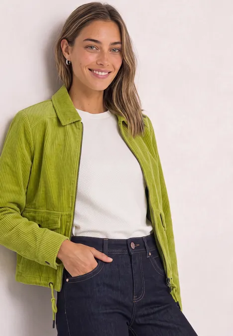 Veste en velours côtelé avec col chemise et fermeture à zip apple lime Junge Frau in grüner Jacke, weißem Shirt und Jeans, die lächelnd in die Kamera blickt.