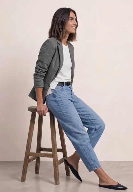 Eenvoudige blazer met reverskraag en patroon Black Frau sitzt entspannt auf einem Hocker, trägt grauen Blazer, weißes Top, blaue Jeans und flache schwarze Schuhe.