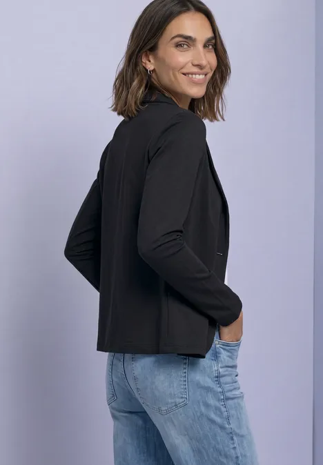 Blazer basique uni Black Eine Frau in schwarzem Blazer und blauen Jeans steht seitlich lächelnd vor hellem Hintergrund.