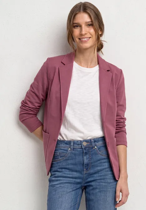 Frau in rosa Blazer, weißem T-Shirt und Jeans lächelt vor hellem Hintergrund.