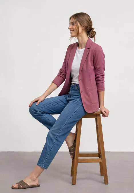 Frau auf Holzhocker, trägt rosa Blazer, weißes Shirt, blaue Jeans und Sandalen.