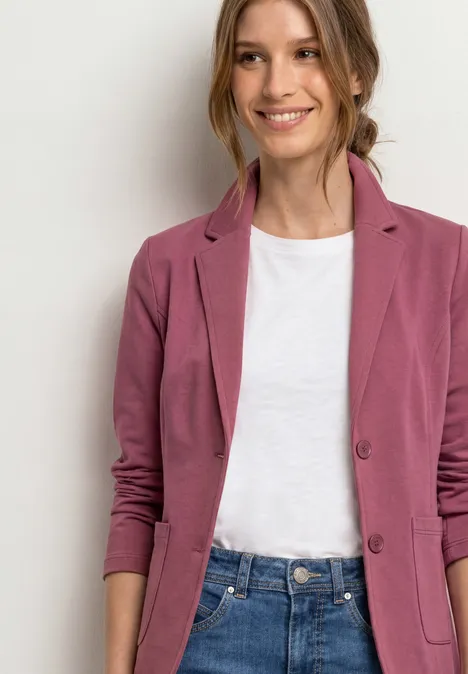 Eine lächelnde Frau in weißem T-Shirt, rosa Blazer und Jeans vor hellem Hintergrund.