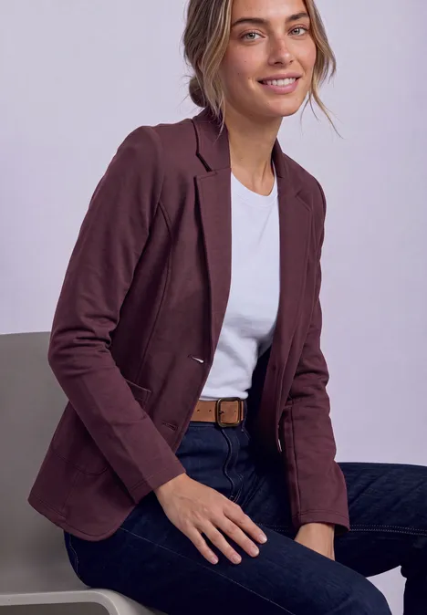 Blazer basique uni rustic rouge Frau in weißem T-Shirt, dunkelrotem Blazer, Jeans und Gürtel sitzt lächelnd.