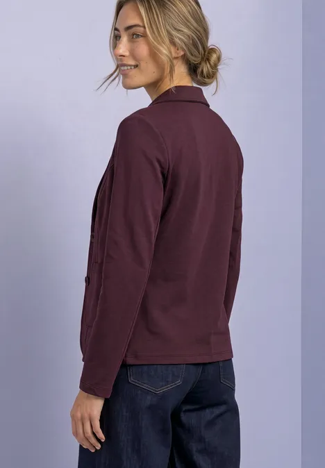 Blazer basique uni rustic rouge Eine Frau mit dunkelrotem Blazer und Jeans blickt über die Schulter, vor lilafarbenem Hintergrund.
