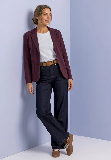 Blazer basique uni rustic rouge Frau in weißem T-Shirt, dunkelblauen Jeans, bordeauxroter Jacke und braunen Schuhen lehnt sich an eine Pastellwand.