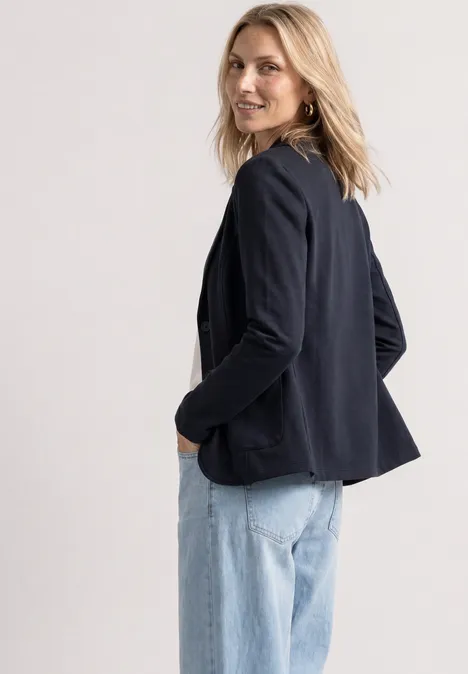 Eine lächelnde Frau trägt eine blaue Jacke und helle Jeans, blickt über ihre Schulter.