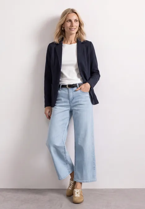 Frau im casual Chic: weiße Bluse, navy Blazer, weite Jeans und Plateau-Schuhe, lächelnd vor hellem Hintergrund.