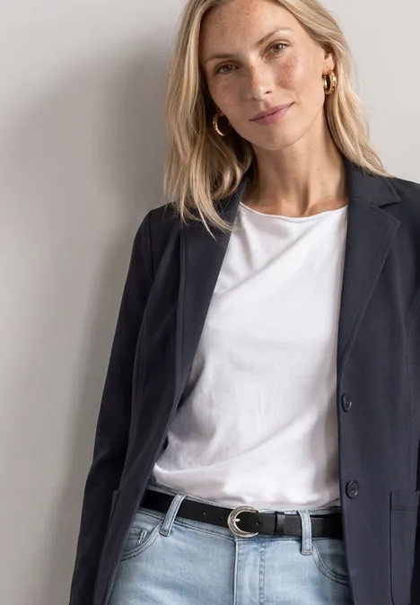 Eine Frau trägt einen schwarzen Blazer, ein weißes T-Shirt und einen schwarzen Gürtel vor hellem Hintergrund.