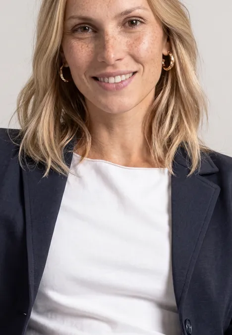 Lächelnde Blondine mit weißen T-Shirt und dunklem Blazer vor neutralem Hintergrund.