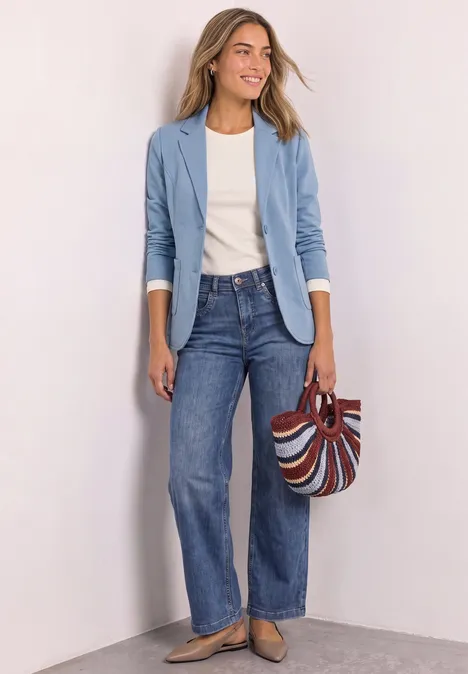Blazer basique uni northern blue mel. Frau in hellblauer Jacke, weißem Shirt, weiten Jeans und gestreifter Tasche vor neutralem Hintergrund.