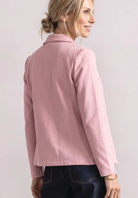 Eine Frau trägt einen rosa Blazer und dunkle Jeans, seitliche Ansicht, sie lächelt.